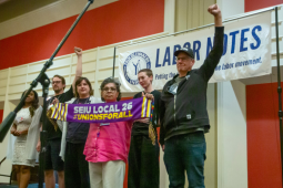 Six workers, of various races and genders, stand together on a ballroom stage with a big "Labor Notes" banner hanging behind them. Three are raising fists in the air, and one holds out a purple "SEIU Local 26, #UnionsForAll" scarf.
