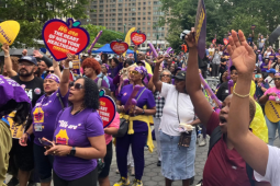 A large crowd of people, mostly Black women, rallies in Manhattan's Foley Square. Many wear purple 1199 T-shirts. Some hold yellow 1199 signs, or red ones shaped like hearts with the message "We are the heart of New York healthcare." The mood is active, with many arms reaching up into the air and mouths opening to speak, chant, or sing.