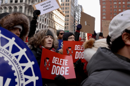 A crowd of people hold red and white signs saying “Delete Doge”