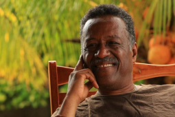 Chest-up photo of Dennis Serrette, a Black man, leaning back in a wooden rocking chair, resting his face gently on one hand, smiling warmly, bathed in warm evening light, with some greenery behind him.