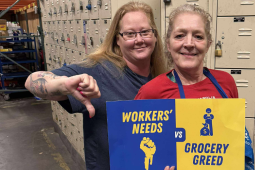 Two white women stand together in front of a wall of lockers, presumably the non-public area of a grocery store, holding a printed blue and yellow sign that reads "Workers' needs vs. grocery greed," with symbols of a fist and Mr. Moneybags. The woman on the left is giving the "thumbs-down" sign. Both smile calmly.