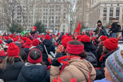 A sea of people, mostly women, of various races, bundled up in winter clothes with red hats and scarves, packs a square in New York City. One person with a megaphone stands elevated a bit in the center, surveying the crowd.