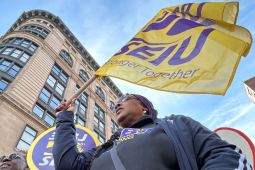 From a heroic low angle, we look up at a Black woman marching in a purple SEIU BJ shirt. The yellow SEIU flag she carries is billowing in the wind, mirroring the curve of the building behind her, against the blue sky. We can see the corners of other picket signs--she is part of a dense crowd.