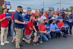 A group of workers in red, blue, or grey shirts hold signs saying “United for a Strong Contract,” and “COLA and Fair Pay Now”  