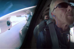 Screenshot of Richard Branson, wearing sunglasses and earphones, in a rocket. Two other crew members are visible behind him. On the left is a window through which you can see part of the outside of the rocket, labeled VIRGIN, and blue sky.