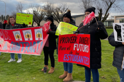 People stand on grass with signs. A red banner says "Seven months without a contract, OMG!" with stylized images of Edward Munch's painting "The Scream." Another sign says "More sick leave" in English, Spanish, and Russian. Another says, in Spanish,"Provide money for the future of students." Another says "Woodburn teachers don't want to strike, but we will." 