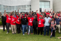 A large group of Black and white women and men stands on grass outside the Mercedes-Benz Training Center, smiling, many with fists in the air. Many wear red T-shirts with a UAW logo and the words "Mercedes Workers United."