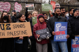 A chanting crowd stands in front of the White House fence. Three people are helping hold a large cardboard sign that says "UE stands with Palestine" with the union's lightning-bolt logo and a slice of watermelon. Other printed signs say "American Postal Workers Union: Fighting for Justice" and "Biden, you are starving Gaza. Permanent ceasefire now!" 