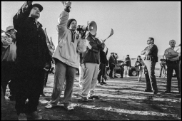 A vintage black-and-white photo shows a line of workers standing outside, one calling into a megaphone, facing down cops with folded arms. They are photographed from a low, dramatic angle.