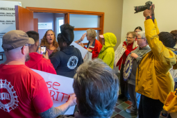 At least a dozen activists crowd up to the door of a VU Manufacturing office, where a company flack is gesuring widely and trying to fend them off. One activist is holding a camera aloft, others holding signs or wearing UAW or Labor Notes gear. A woman 