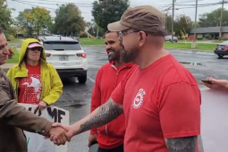 Screenshot shows Israel Cervantes on the left, in a weathered brown leather jacket, shaking hands with Sean Crawford, right, in a red UAW T-shirt and a baseball cap, while a few other activists carrying handmade signs look on approvingly. They're standing in a parking lot after a rain.