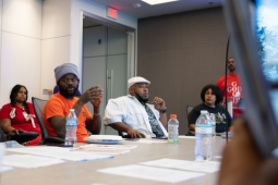 Several Black workers, women and men, sit around a conference table. In the middle of the frame, a man in an orange T-shirt is talking, looking around at the group; others are listening thoughtfully. Everyone's expression is serious.