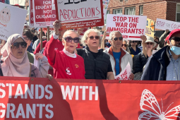 Workers, mostly women, carry a big red banner with white letters: "PSC stands with immigrants." They are in a big crowd. Some also carry picket signs, some printed and some handwritten, with messages (some partially visible) including "Dictators don't belong here," "America? Fight for it!" "Oranges don't belong in the White House," "Stop the war on immigrants," and "Stop the coup." One woman wears a hijab.