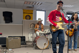 A band performs in a union hall. The guitarist in front is a tall man in a red T-shirt with a design that starts "No Power" (his guitar blocks the rest of hte slogan" and the arm says "Stronger." There's a woman on bass, a man in a cowboy hat on drums, and a man holding an accordion sitting at a keyboard. Behind them is a yellow AFL-CIO banner. For some reason a black T-shirt is draped over a framed item on the wall.