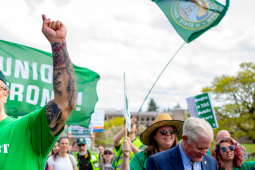 A man in a backwards green ballcap and an AFSCME green T-shirt printed with the word "Activist" raises his heavily tattooed arm up to the sky. Behind him other marchers are visible, crossing the capitol campus, some with signs (not readable) and one waving a Washington state flag on a tall pole.