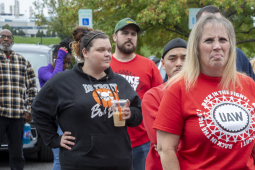 A group of autoworkers stand around outside looking at the camera--the closest is a blonde woman with a red shirt that says “UAW Back in the Fight.”