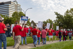 Workers in red shirts picket outside. Many of the picket signs are white with UAW logo, Local 412, and the words "Stellantis Bargaining in Bad Faith."