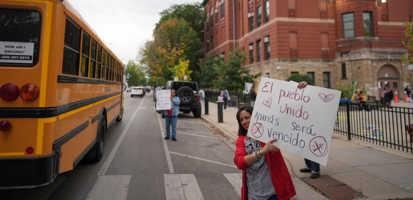 A teacher holds a sign between a school bus and the front of a school, with students visible in the background