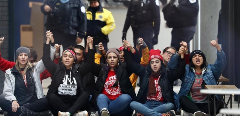 Teachers seated on sidewalk with joined hands raised; police standing behind them