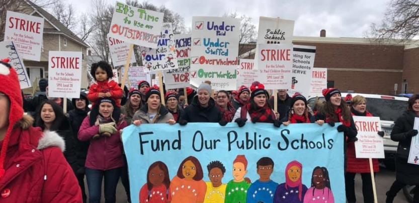Strikers carry a banner "Fund Our Public Schools" with a rainbow of faces. Picket signs say "Strike" and "It's Time to Use Our Outside Voice." One kid is carried on an adult's shoulders.