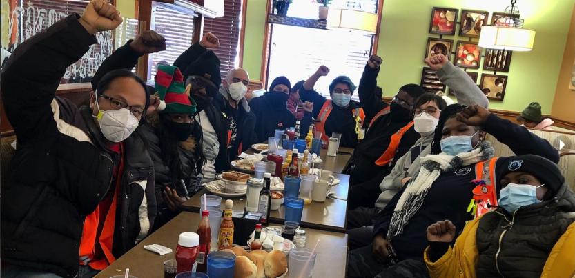 A group of workers sitting at a long restaurant table, some in safety orange vests and wearing surgical masks, raise their fists and look at the camera. 
