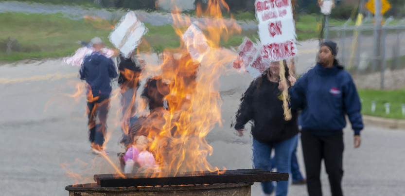 Strikers march holding picket signs that read "On Strike," while a fire rises from a burn barrel to keep them warm.