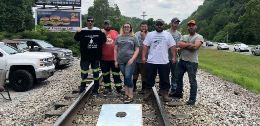 Five people stand on railroad tracks, with a cornhole game at their feet. Some wear "pay us what we are owed" shirts.