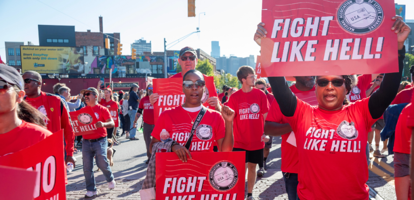 Black and white women and men march through a Detroit street. Everyone is wearing a red "fight like hell" T-shirt with the NALC logo, and some people are carrying printed signs with the same design. Most prominent in the frame, looking directly into the camera, are two Black women in sunglasses, one raising a fist at shoulder level and the other holding a sign above her head. 