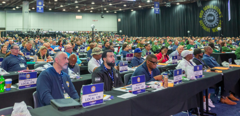 UAW delegates (men and women, Black and white) sit listening at the union's 2026 convention. Table signs identify those in the foreground as from Region 8, Locals 129 and 276.