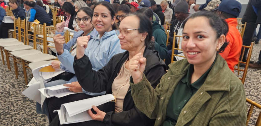 Four women of a range of ages, seated in a row, part of a bigger crowd in what looks like a hotel ballroom, smile and raise fists. Each holds in her lap a stapled packet of printed pages, perhaps a summary of a tentative agreement.