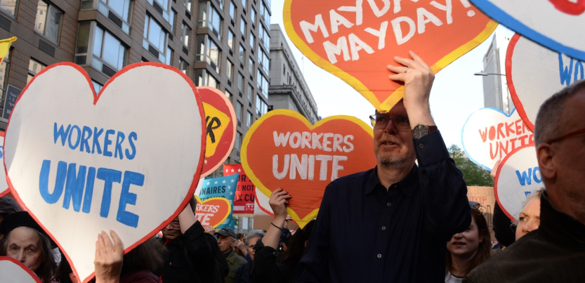 People march with heart-shaped signs saying “workers unite” and “May Day!”