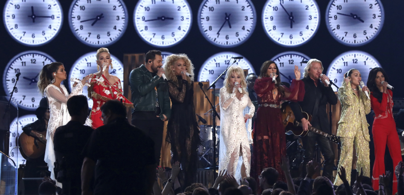 A row of musicians with large clocks behind them perform, with Dolly Parton in the center