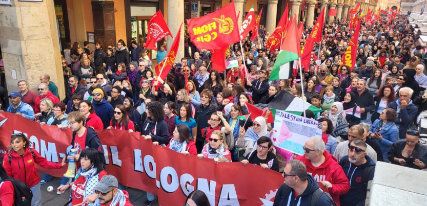 View of hundreds of people, a section of a giant march, filling an Italian street. Some marchers carry a huge red banner "FIOM-CGIL Bologna," some carry union flags or Palestinian flags, and one carries a handmade sign that says "Palestina Libera" with the flag and hearts. Mouths are open to sing or chant. Picturesque columns along the street. Marchers are diverse in race and gender.