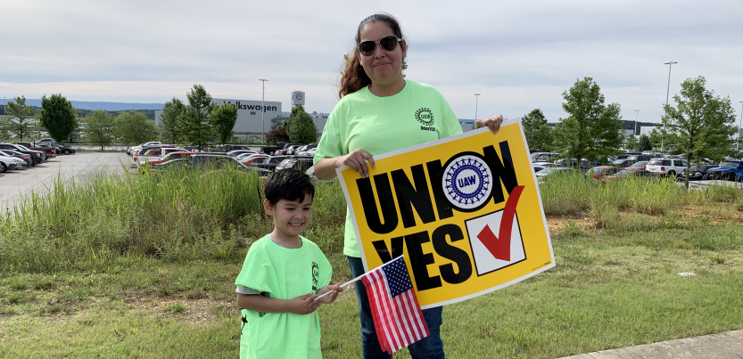 Woman holding "UNION YES" sign and child holding U.S. flag, with VW plant behind