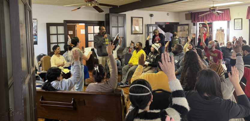 A group of people in chairs crowd a room listening to a speaker.
