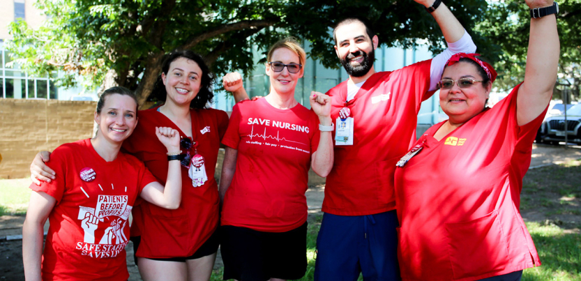 A group of five nurses in red shirts pose with their fists raised.
