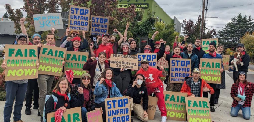 A group of workers and supporters hold cardboard signs reading Stop Unfair Labor Practices and Fair Contract Now. Many are wearing red bandanas.