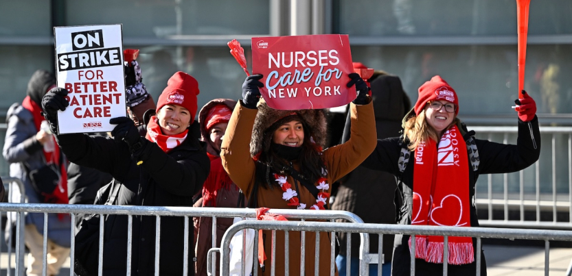 Three smiling nurses stand against a barrier with signs about patient care