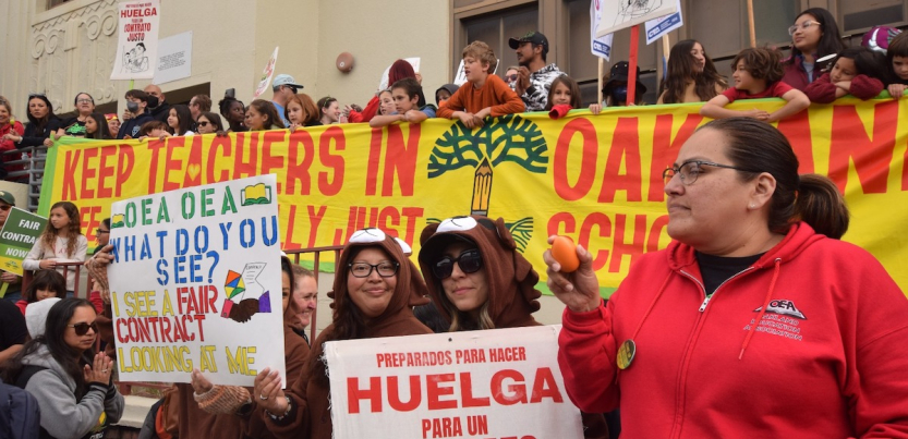 A crowd of adults and children hold a banner saying Keep Teachers in Oakland Schools while other signs say Huelga para un Contracto Justo and Fair Contract Now.