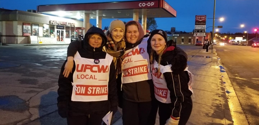 Four smiling women wearing "UFCW Local ON STRIKE" signs stand in front of a Co-op gas station, arms around each other's shoulders