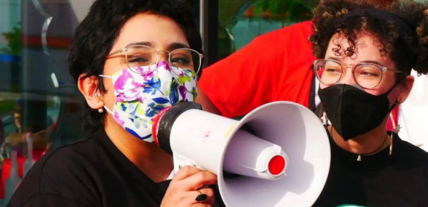 One masked worker speaks into a bullhorn as another looks on. Both look happy and young.