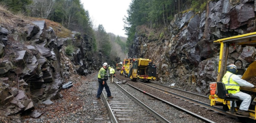 Workers in reflective yellow jackets work outdoors on railroad tracks, with heavy machinery, surrounded by rocky cliffs and forest