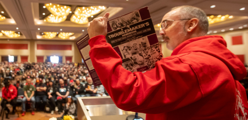 Seen from the side/back, Shawn Fain in a red hoodie holds up a battered purple copy of A Troublemaker's Handbook. Arrayed in front of him can be seen a huge seated crowd in the ballroom for the closing plenary.