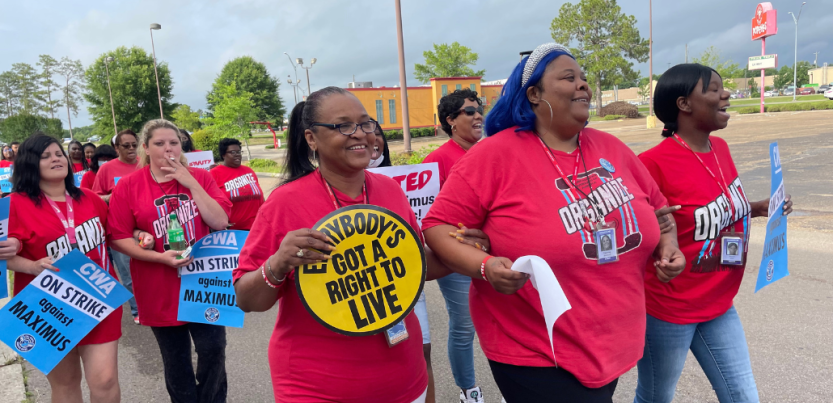 Women, mostly Black women, march outdoors in red T-shirts that say "Organize" with a telephone logo. Most prominent in the foreground are three women who have linked elbows and are smiling big. The woman on the left has a yellow round sign that says "Everybody's got a right to live." The woman in the middle has blue hair. The woman on the right is holding a light blue sign we can't see, but it probably matches signs visible behind which say "CWA on strike against Maximus." One woman is smoking.