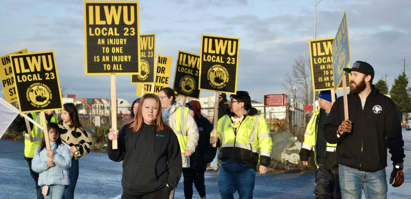A group of union members and supporters march holding black and yellow ILWU Local 23 and Unfair Labor Practice signs.