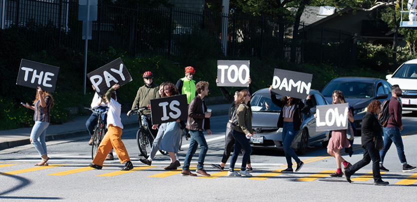grad students blocking traffic in the street with signs that read "The Pay Is Too Damn Low"