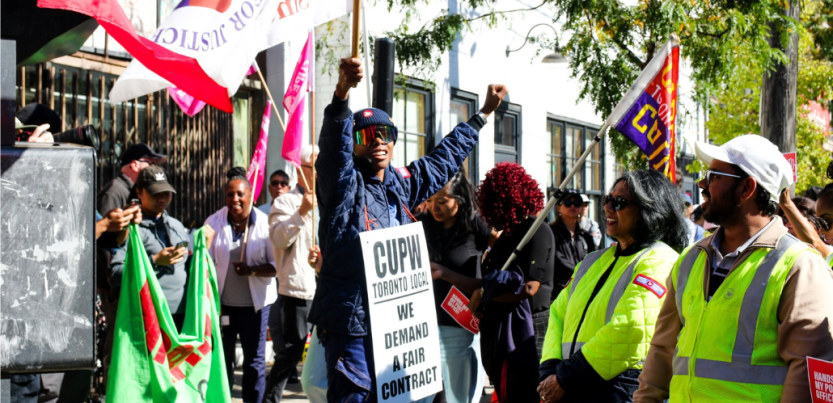A Black worker in postal uniform, wearing reflective shades and a sandwich board sign that says "CUPW Toronto Local, We demand a fair contract," raises one fist in the air and a flag on a stick in the other hand. The flag is only partially visible but includes the word "Justice." This person is standing up on something we can't see, a step or box. Smiling people around them are holding various banners and signs, some wear safety yellow. Variety in race and gender. In sunlight dappled by shade of tree. 