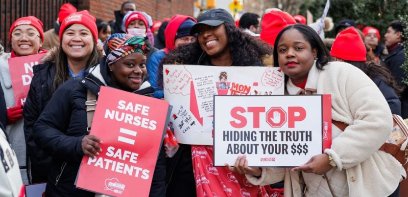 Three Black women nurses pose for camera; two are smiling, one looks determined. Two hold printed NYSNA signs: "Safe nurses = safe patients" and "STOP hiding the truth about your $$$" and the other holds a handwritten sign, partly covered, long text. Lots of red NYSNA hats are visible in the dense, upbeat crowd behind them.