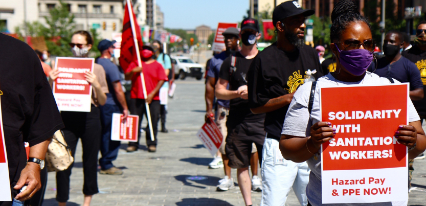 Masked people stand outside holding red printed signs. Visible sign in foreground says: "Solidarity with Sanitation Workers! Hazard Pay and PPE Now!"