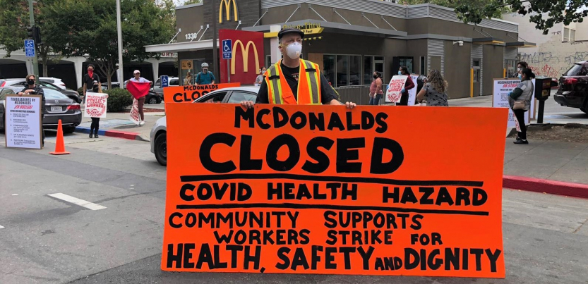 masked worker in front of mcdonald's holds big orange sign: "MCDONALD'S CLOSED, COVID HEALTH HAZARD, COMMUNITY SUPPORTS WORKERS STRIKE FOR HEALTH, SAFETY AND DIGNITY"